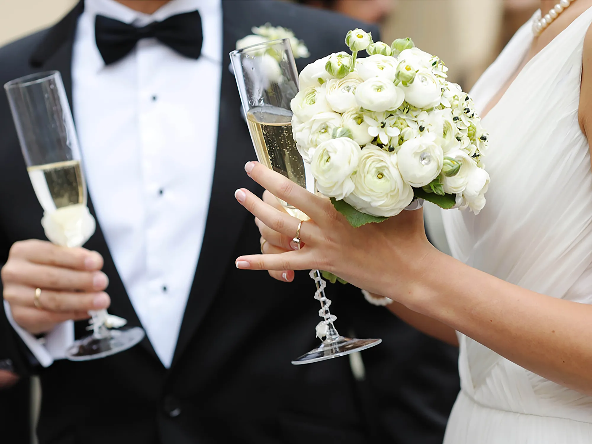 Nell'immagine, il primo piano sulle mani di una coppia di sposi: entrambi reggono un calice di spumante e la sposa ha in mano anche un bouquet di fiori bianchi.