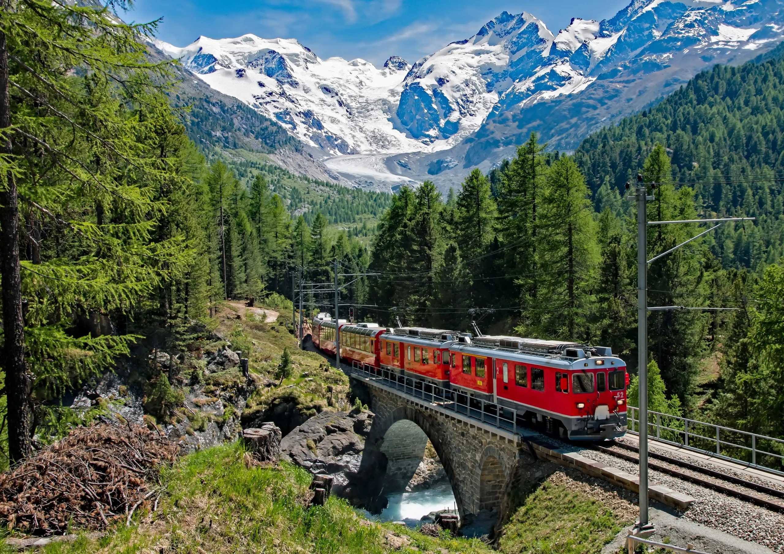 Nell'immagine, il trenino rosso del Bernina che attraversa un ponte, circondato da alberi e con le montagne innevate sullo sfondo.