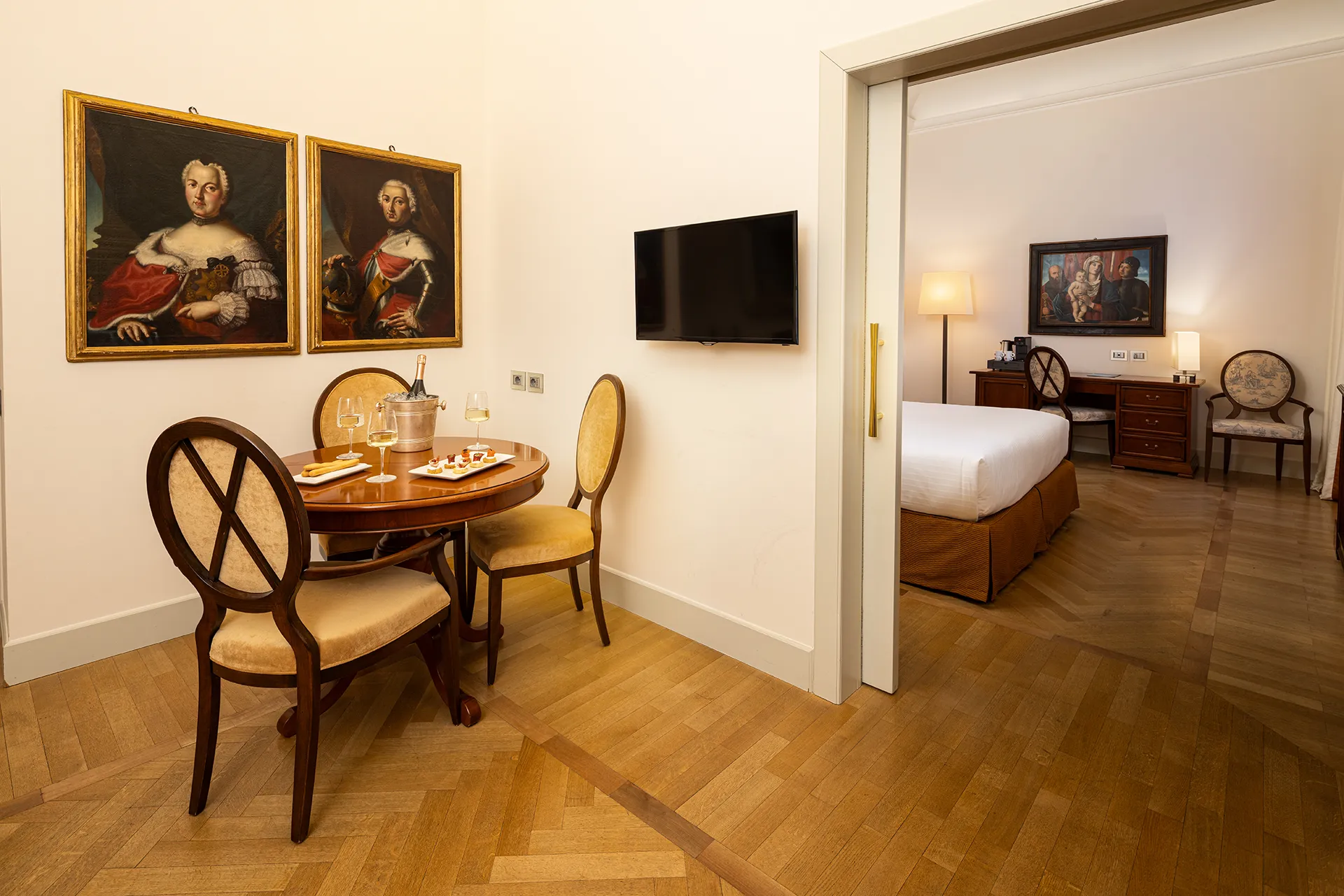 Nell'immagine, la zona giorno della Suite del Grand Hotel della Posta: si vede un tavolo tondo apparecchiato con delle tartine e una bottiglia di spumante. Alla parete, sulla sinistra sono appesi due quadri e sulla destra la TV.