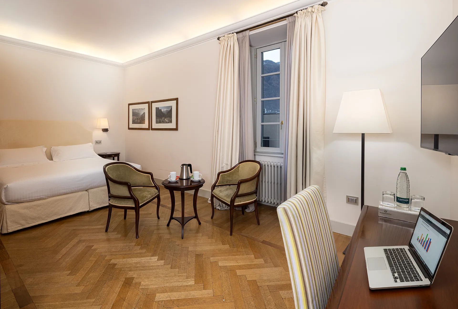 Nell'immagine, la camera Superior del Grand Hotel della Posta: si vede il letto matrimoniale e, ai piedi del letto, un tavolino tondo con due poltroncine. Sul tavolo è poggiato un bollitore con due tazze. Sulla destra, si intravede un mobile su cui è poggiato un laptop acceso e la TV appesa alla parete.