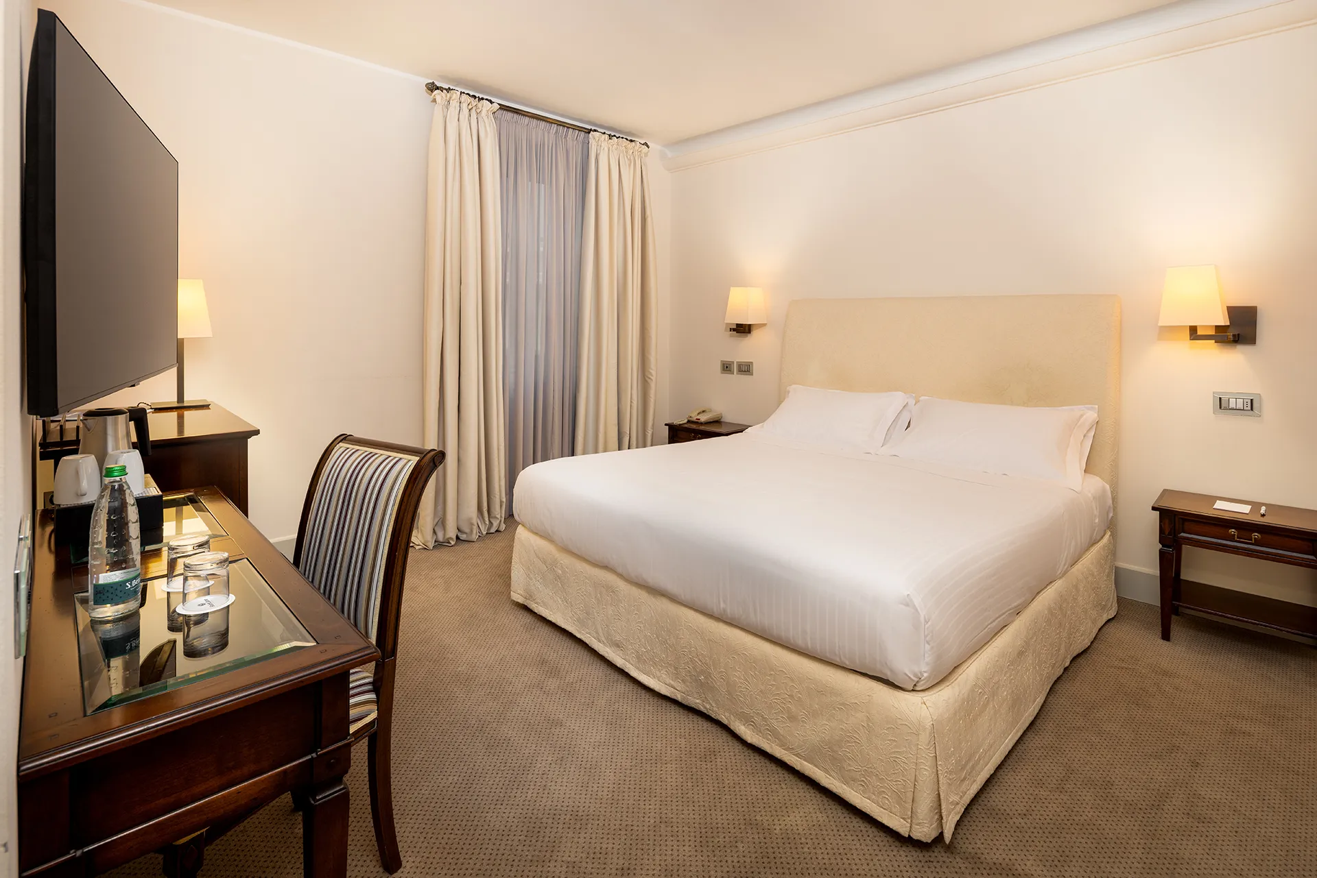 Nell'immagine, la camera Classic del Grand Hotel della Posta: si vede il letto matrimoniale e, di fronte, un mobile su cui sono poggiati un bollitore con due tazze e una bottiglia d'acqua con due bicchieri di vetro. Alla parete è appesa la TV.